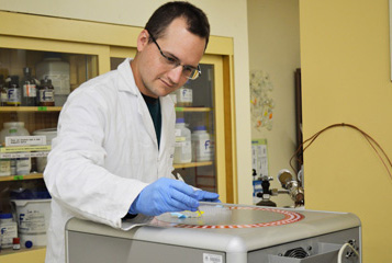 Researcher in lab with lab coat and gloves on, reaching across a square machine with holes along the top to hold vials