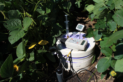 Tucked among rows of mature soy plants is a white bucket-shaped device with a temperature display screen, keypad, injection syringe, and tubing. The black flexible tubing connects to a smaller gray cylinder sunk into the soil nearby.