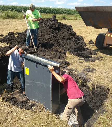 Two people are lowering a gray metal box into a trench of dark soil. A third person is in the background, along with a bulldozer. on each side of the trench is tan grass from last year's growth and beyond them a field of corn.