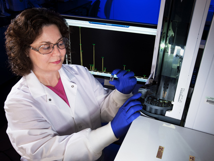 A scientist wearing  lab coat, eye protection, and blue gloves inserts a small vial of clear liquid into a chromatograph. Behind her is a computer screen showing a chart.