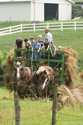 Two horses pull a wagon full of straw with several kids on top. A farmer in a broad-rimmed hat watches as a child holds the reins. In the background is a white fence for the pasture and a white barn with a door open.